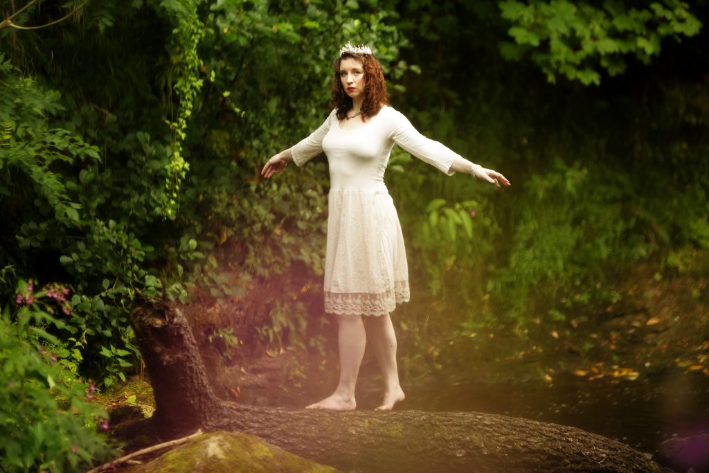 photo of author maya macgregor walking barefoot across a log above a river with their arms outstretched, wearing a soft, knee-length ivory dress and a sparkling tiara settled on loose auburn curls